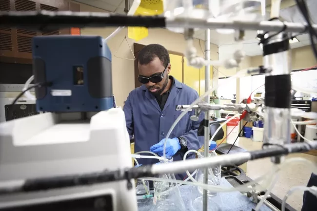 Person in a lab wearing sunglasses, surrounded by beakers, tubes and other lab equipment.